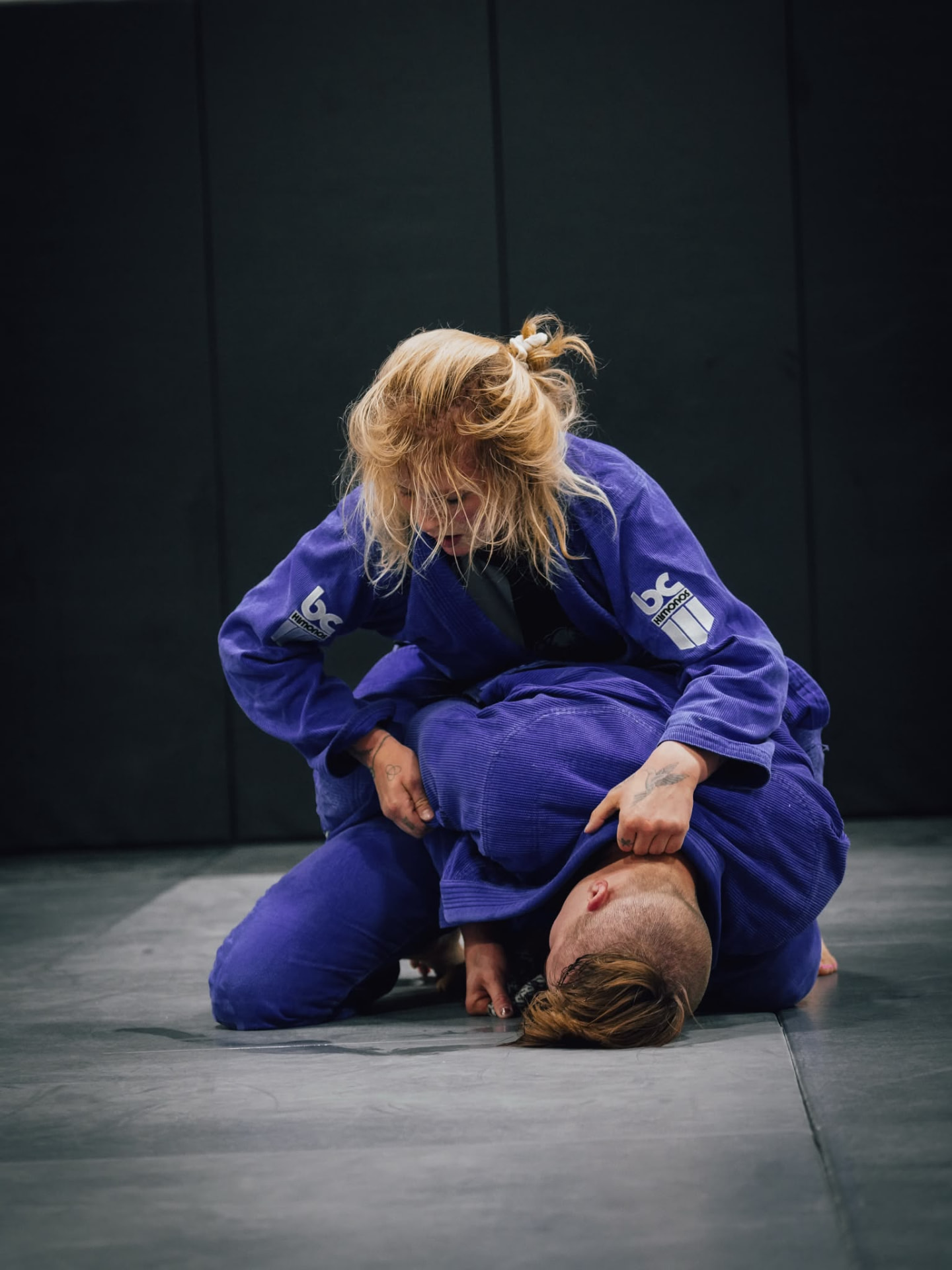 Chayse in a blue gi driving forward over a training partner on the mat, controlling his lapel and sleeve
